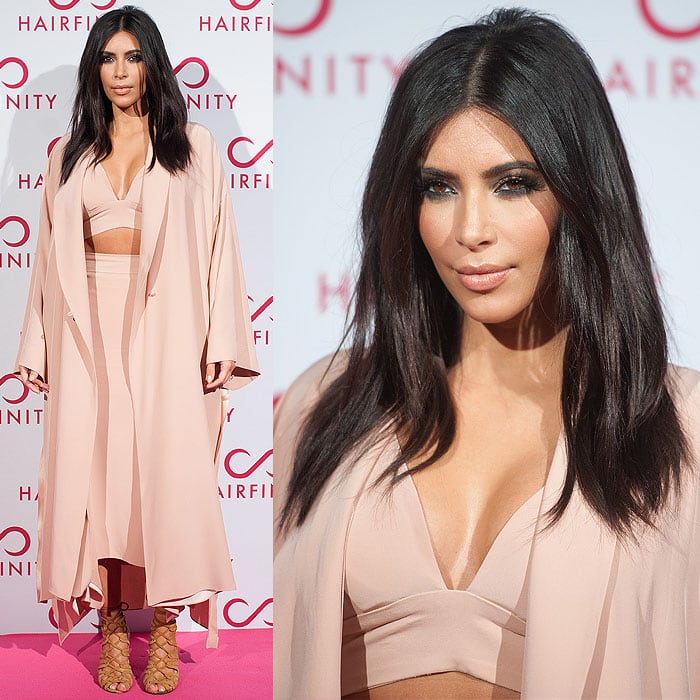 Kim Kardashian working her signature stance in a pale pink bralet, a midi skirt, a drapey overcoat, and Hermès lace-up sandals