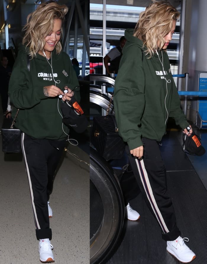 Rita Ora wearing a dark green hoodie, black trackpants, and white Reebok "Classic Leather" sneakers at LAX