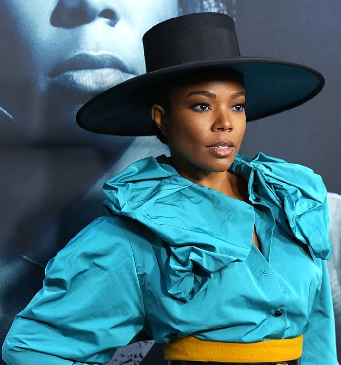 Gabrielle Union wearing blue eyeliner, a top hat, a blue ruffled blouse, and yellow pants.