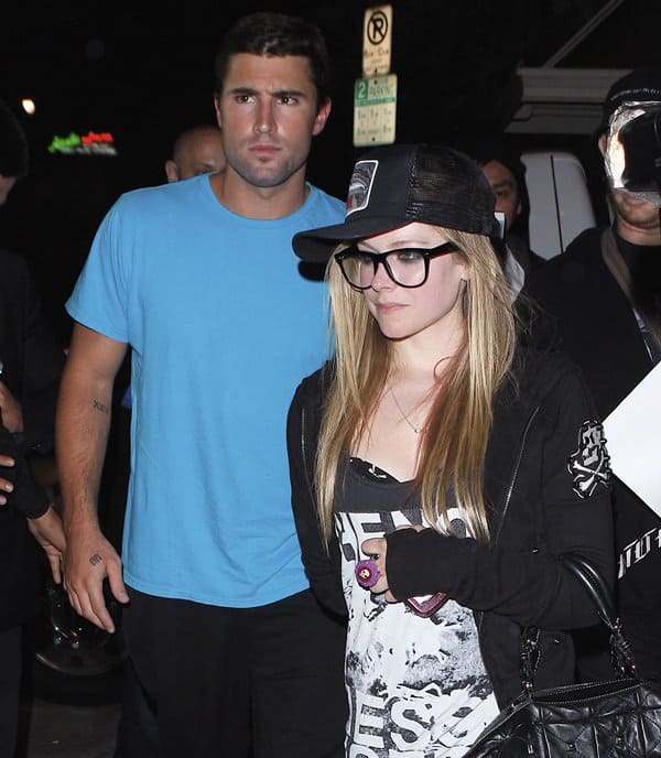 Avril Lavigne leans into her edgy street style with oversized glasses, a trucker hat, and graphic layers while stepping out hand-in-hand with Brody Jenner, who keeps it casual in a bright blue tee