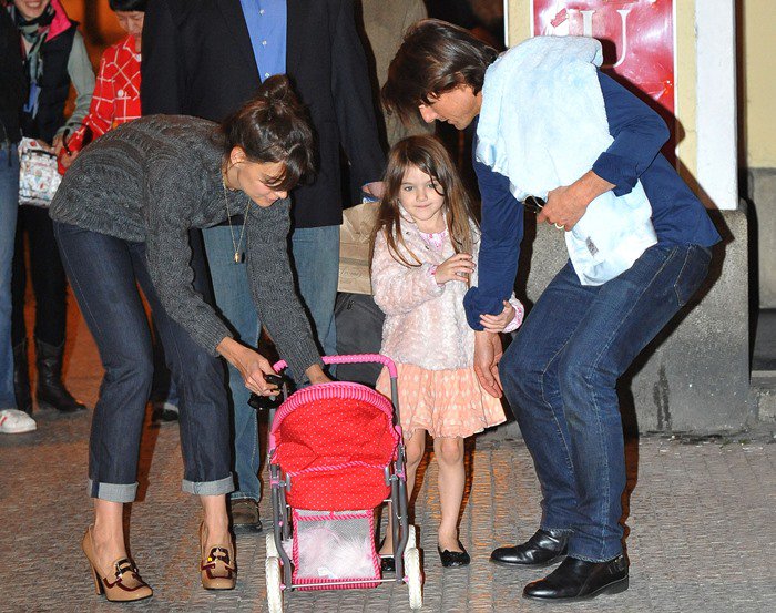 Katie Holmes, dressed in a cozy gray sweater and Prada loafers, shares a sweet family moment in Prague, helping her daughter Suri with a toy stroller as Tom Cruise looks on