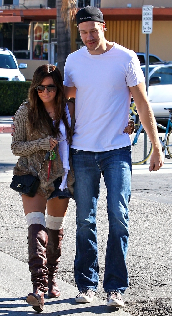 Ashley Tisdale exudes cozy chic in a fur-trimmed cardigan, denim shorts, thigh-high socks, and slouchy brown boots, perfectly contrasted by Scott Speer's casual white tee, jeans, and sneakers