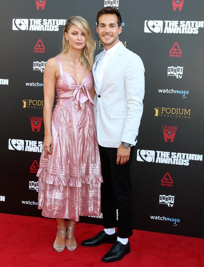 Melissa Benoist dazzles in a metallic pink tiered dress with strappy silver heels, posing alongside husband Chris Wood in a crisp white blazer at the 45th Annual Saturn Awards