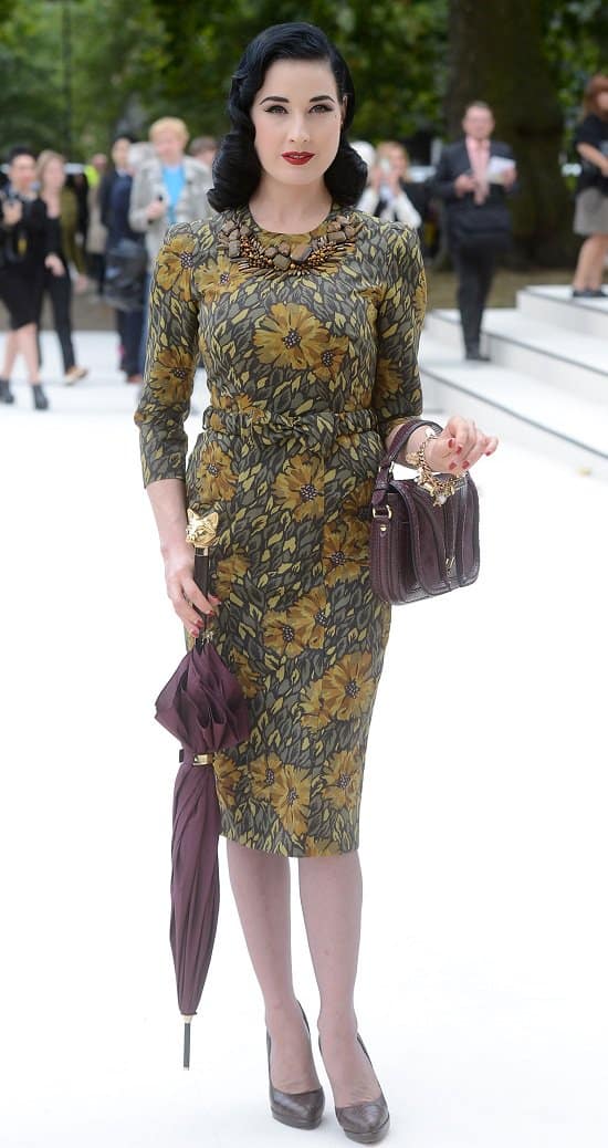 Fashion icon Dita von Teese elegantly carries a whimsical cat-detailed maroon umbrella, perfectly complementing her retro 50s vintage floral dress, at the London Fashion Week's Burberry arrivals
