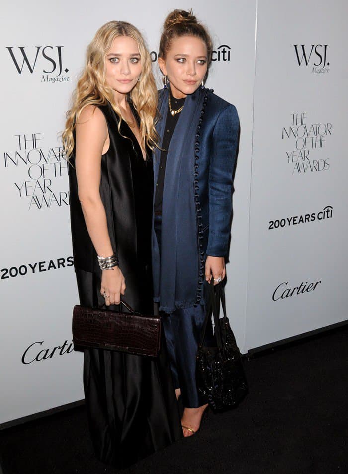 At the Museum of Modern Art in New York, Mary-Kate Olsen, draped in a tailored blue silky suit, and Ashley Olsen, in a deep-V-neck black maxi dress, gracefully pose at the WSJ Magazine's Innovator of the Year Awards, exuding a blend of modern sophistication and avant-garde style