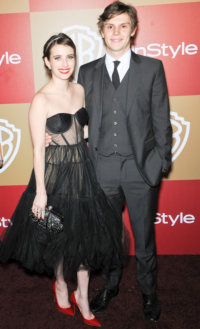 Evan Peters looks sharp in a tailored gray three-piece suit with polished black dress shoes, standing slightly taller than Emma Roberts in her dramatic black Maria Lucia Hohan dress and red Christian Louboutin heels