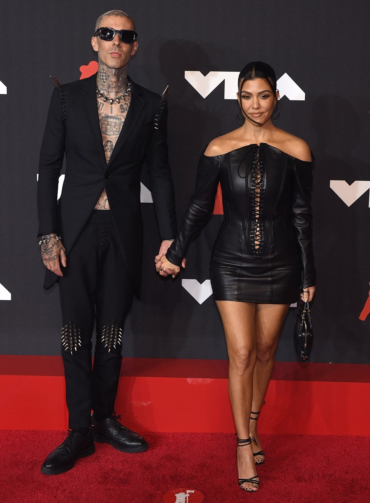 Kourtney Kardashian and Travis Barker make a bold style statement at the 2021 MTV Video Music Awards, with Kourtney in a sultry lace-up leather mini dress and Travis in an edgy spiked suit, exuding rockstar romance on the red carpet