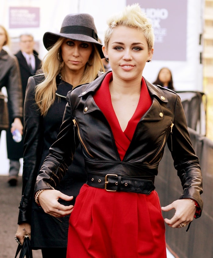 Miley Cyrus elevates her fashion game by pairing a fiery red Rachel Zoe ‘Edith II’ jumpsuit with a chic Moschino leather moto jacket, demonstrating a perfect blend of elegance and rebel chic