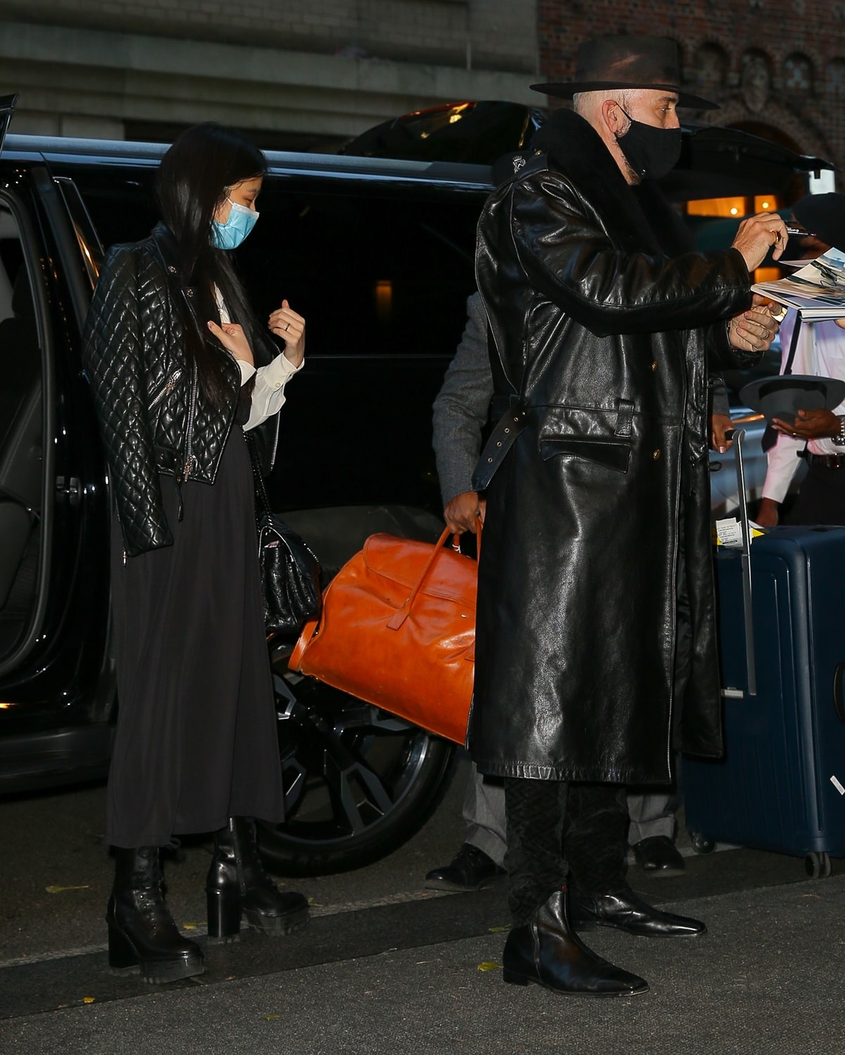 Nicolas Cage and his wife, Riko Shibata, arrive in style at their New York City hotel on November 27, 2021, with Riko rocking a chic quilted jacket, a flowing black skirt, and platform combat boots, while Cage opts for a dramatic leather trench coat paired with sleek pointed-toe boots