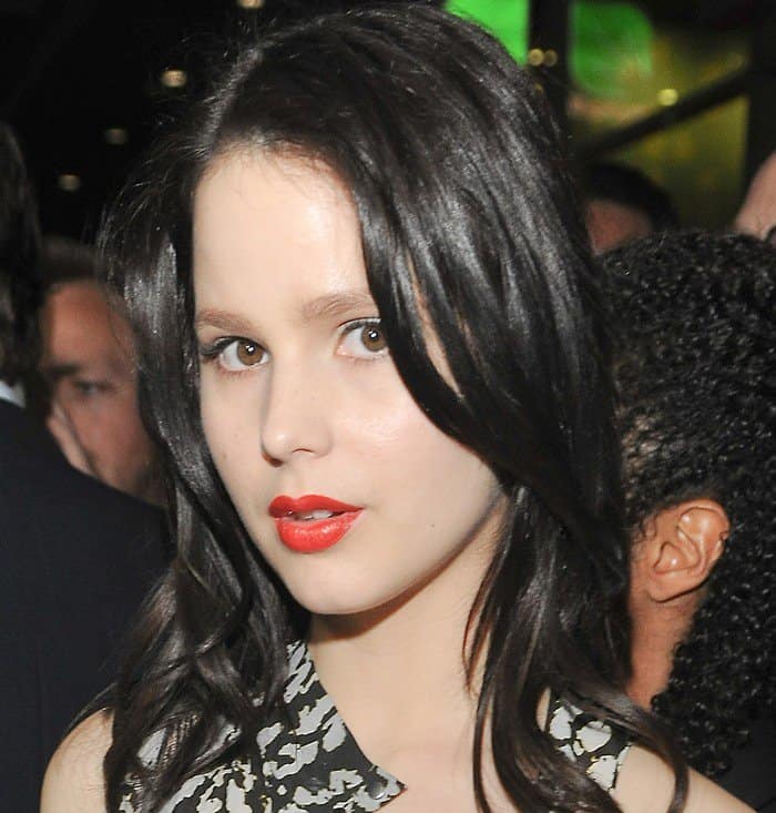 Rachel Korine's makeup complements the dress with a classic bold red lip, and she sports a simple, straight hairstyle with a side part