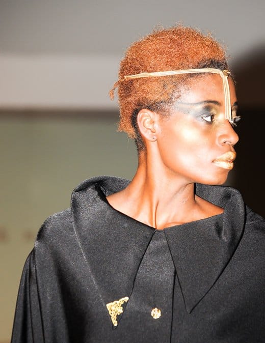 Striking Profile at Birmingham Fashion Week 2013: A model showcases minimalist sophistication with a sleek black coat accented by a delicate golden headpiece