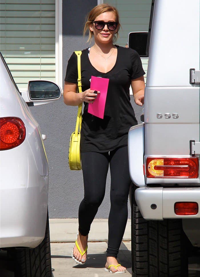 Captured on May 24, 2013, Hilary Duff leaves her Pilates class in Toluca Lake, dressed casually in American Apparel leggings, a black Lanston tee, accessorized with Wildfox sunglasses, a vibrant Rebecca Minkoff 'Skylar' bag, and Havaianas flip-flops