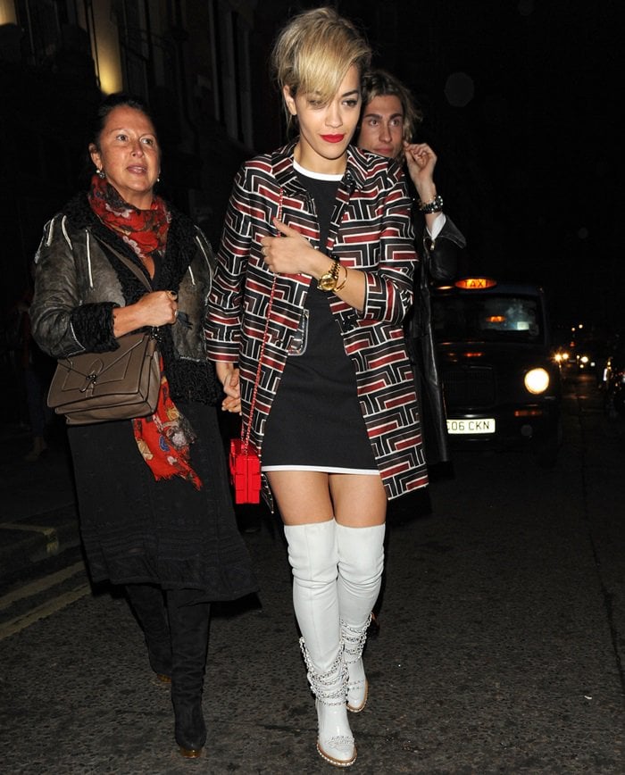 Rita Ora wearing a printed coat, an LBD, white over-the-knee boots, and a striking red box clutch with intertwining Cs on it