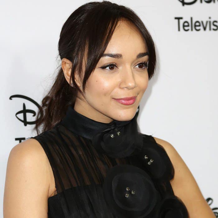 Ashley Madekwe radiates elegance in a sheer black dress embellished with oversized floral accents, complemented by a chic, high-neck collar and soft, natural makeup