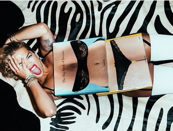 Rita Ora playfully strikes a pose on a zebra-striped rug, tongue out and book in hand, capturing a moment of cheeky fun