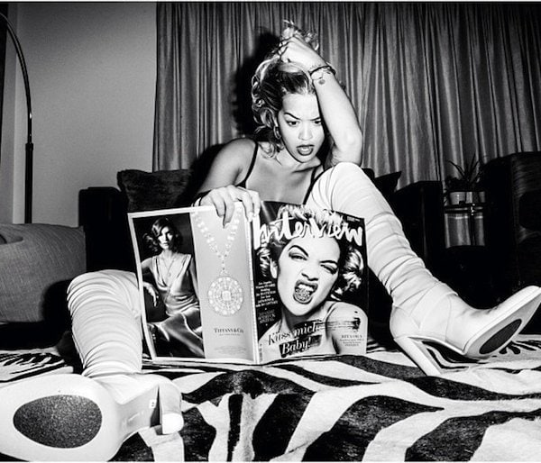 Rita Ora exudes retro glamour while casually lounging with a magazine, sporting striking white thigh-high boots and a classic pin-up hairstyle