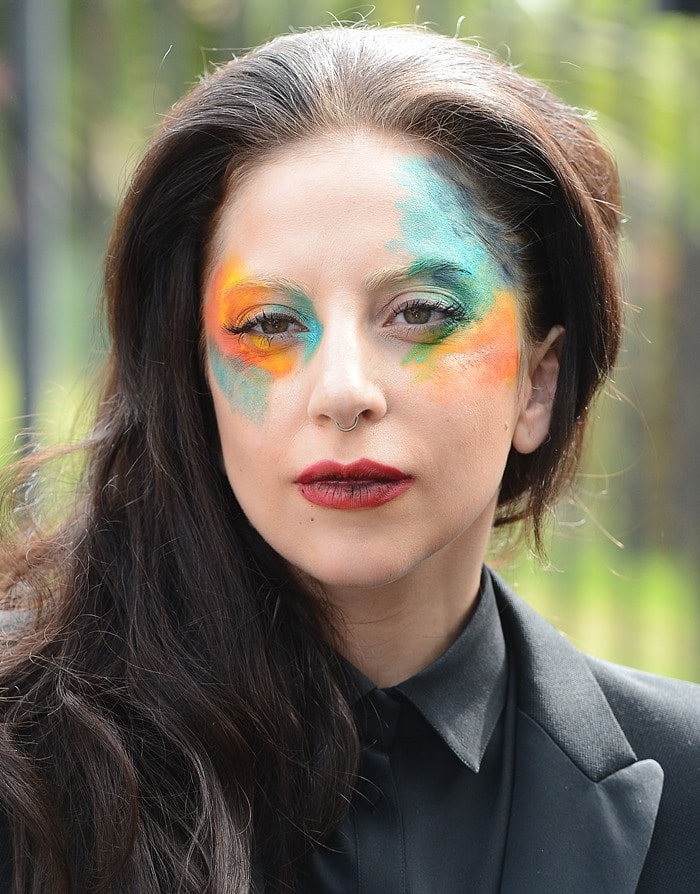 Lady Gaga sports a vibrant, multicolored makeup look, combining bold orange, green, and blue shades on her cheeks, complementing her dark, tailored outfit and subtle jewelry