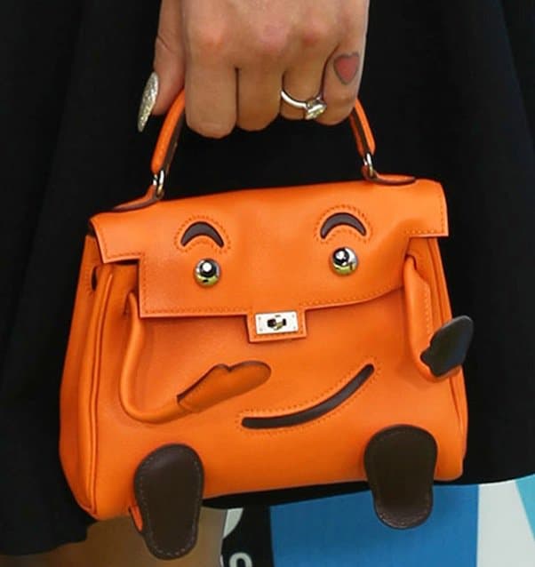 Detailing her playful side, Kelly Osbourne complements her sophisticated black dress with a charming vintage orange Hermès handbag featuring a smiley face, pictured here alongside her engagement ring from fiancé Matthew Mosshart