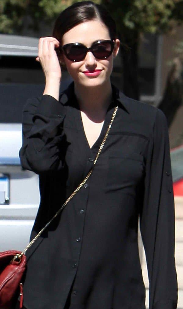 Emmy Rossum looks effortlessly chic in a black button-down top and cat-eye sunglasses, accessorized with a red Zadig & Voltaire pochette and coral-red lips