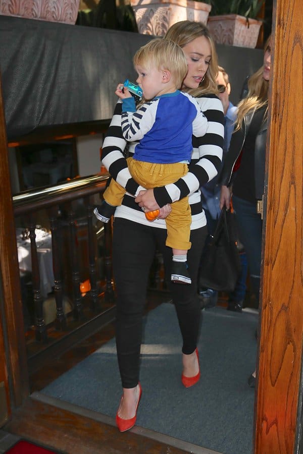Hilary Duff pairs a black and white striped top with black pants and red heels, adding a pop of color while carrying her child
