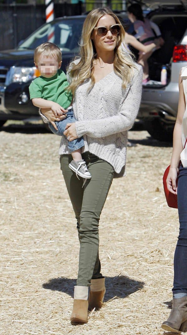 Kristin Cavallari looks effortlessly chic in green rolled-up pants, a cozy knit sweater, and tan ankle boots while carrying her child
