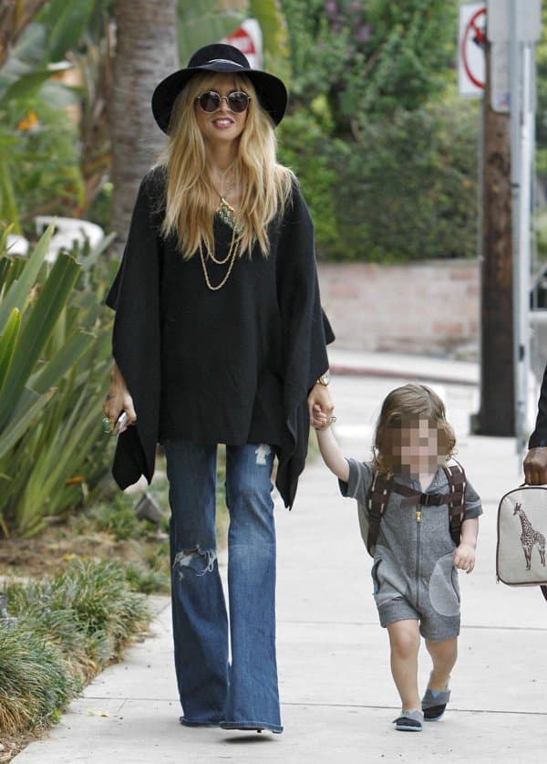 Rachel Zoe embraces boho chic with a wide-brimmed hat, black poncho, and distressed flared jeans while walking with her child