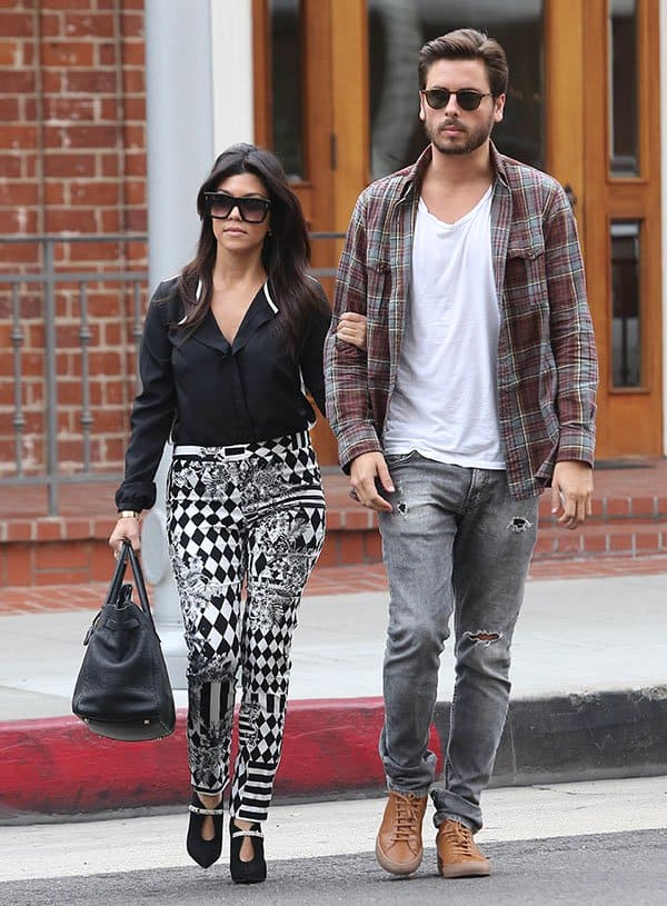 Kourtney Kardashian (5′ 0″) stuns in a chic black blouse and patterned pants, while Scott Disick (5′ 11″) rocks a casual look with torn jeans and a plaid shirt, highlighting their noticeable height difference