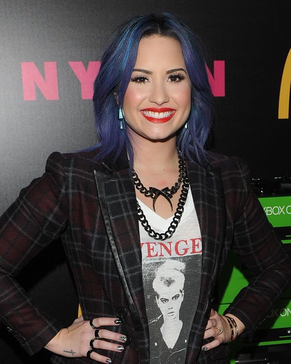 Demi Lovato wearing a Marissa Webb plaid jacket over a graphic tee, accessorized with bold jewelry and a bright red lip