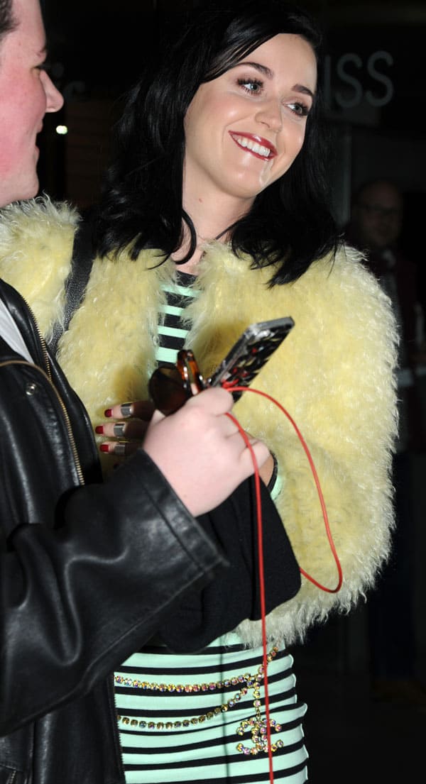 Katy Perry in a mint-and-black striped dress with a yellow fur jacket, smiling while signing autographs