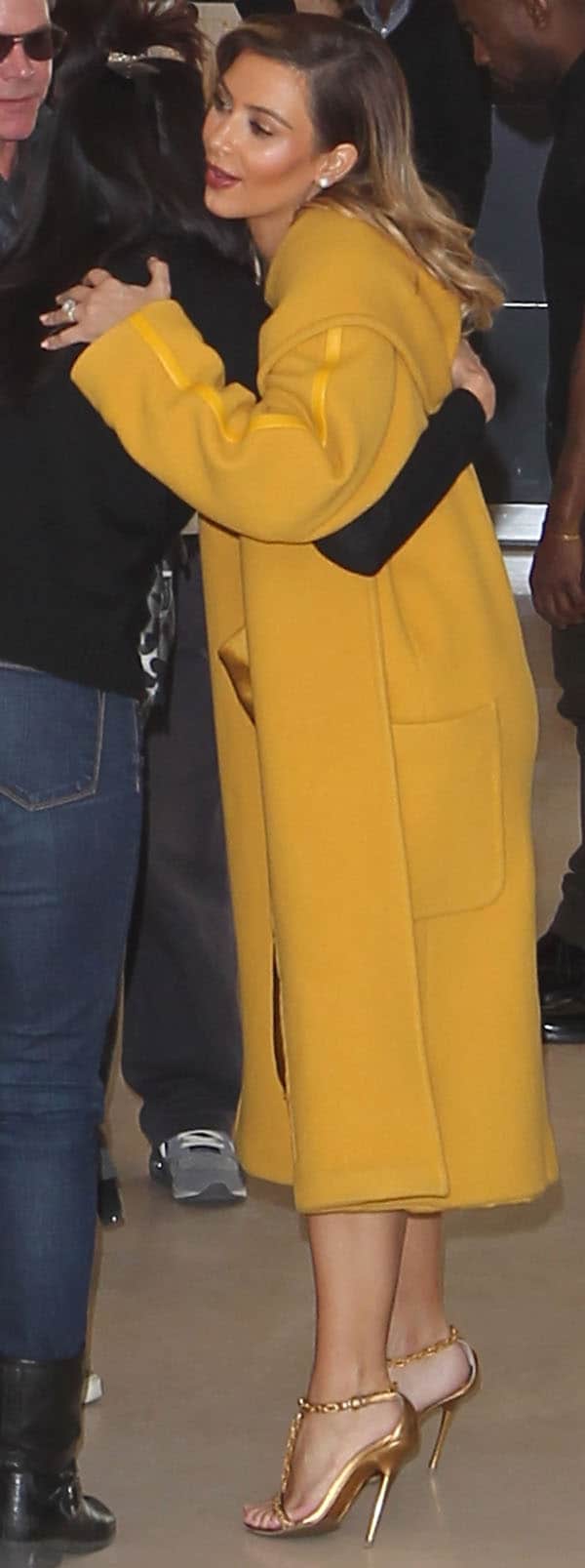 Kim Kardashian greets a friend warmly while flaunting her chic style in a mustard yellow coat and matching silk dress, completed with elegant gold chain-link heels