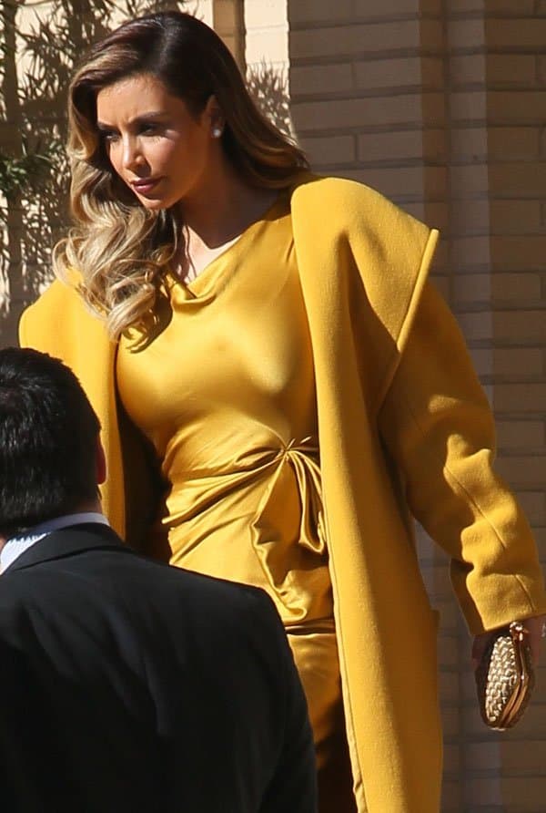 Kim Kardashian stuns in a mustard yellow silk dress paired with a matching coat, accessorized with a gold clutch and her signature voluminous waves, exuding elegance and style