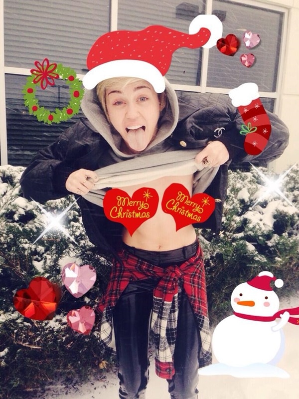Miley Cyrus spreading holiday cheer in a festive and playful photo, lifting her shirt to reveal "Merry Christmas" heart pasties while wearing a Santa hat and surrounded by Christmas-themed decorations