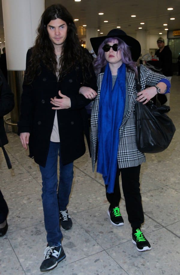 Kelly Osbourne and fiancé Matthew Mosshart make a stylish entrance at Heathrow Airport, with Kelly sporting a chic houndstooth coat, vibrant blue scarf, and a trendy floppy-brimmed hat