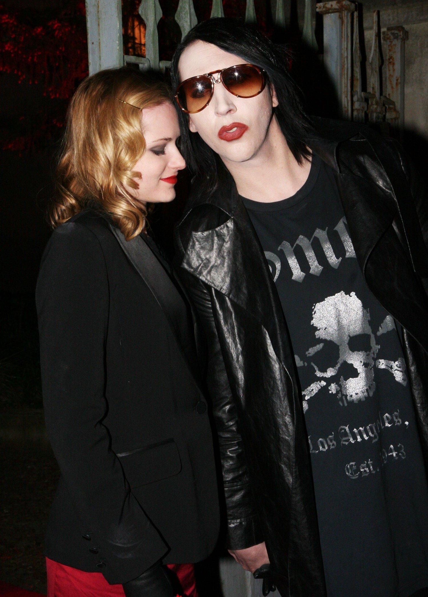 Marilyn Manson and girlfriend Evan Rachel Wood coordinate in edgy all-black ensembles, with Evan adding a bold pop of color in crimson pants and Manson opting for a skull-print tee and leather trench