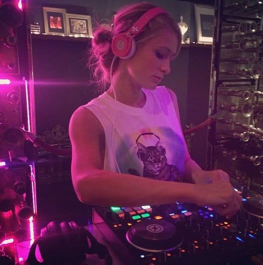 Paris Hilton focuses on her DJ set, sporting a casual look with a playful cat-themed tank top and pink headphones, bringing energy to the decks