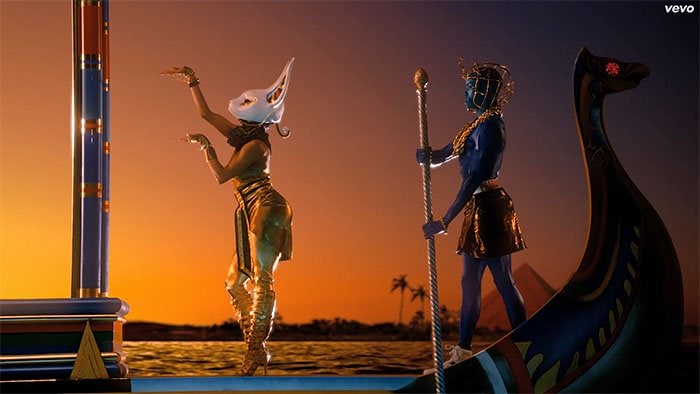 A backup dancer stuns in knee-high gladiator sandals and a gold ensemble while escorting a vibrant blue-skinned figure