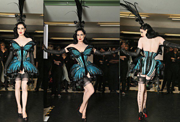 Dita Von Teese dazzles in a dramatic turquoise-and-black butterfly corset, complete with sheer stockings, leather gloves, and a striking feathered headpiece, embodying retro glamour at its finest