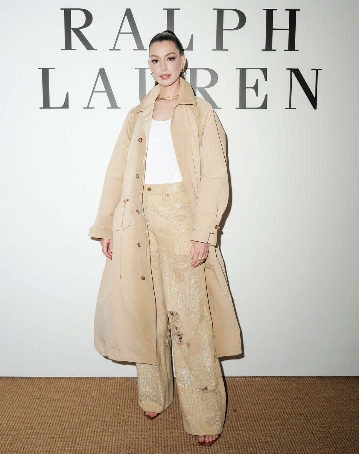 Anne Hathaway brings effortless sophistication to the Ralph Lauren Spring 2025 show on April 17, 2025, in New York City, wearing a sand-toned trench coat layered over sequined wide-leg pants and Gianvito Rossi Ribbon stiletto sandals, styled with gold Bvlgari Serpenti jewelry and accessories