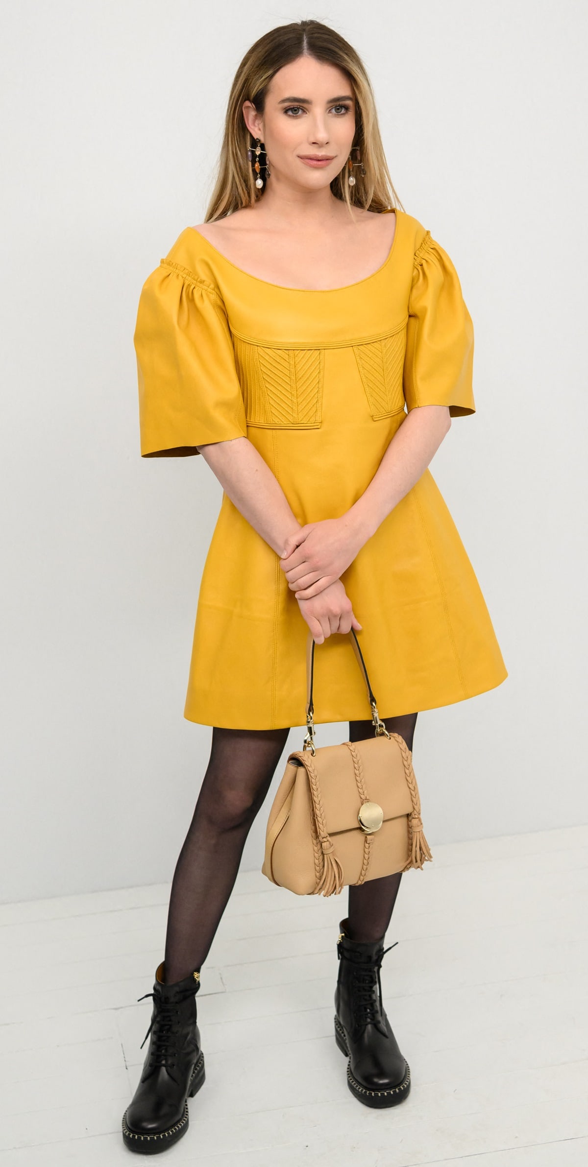 Emma Roberts attended Chloé's fall 2023 show on March 2, 2023, during Paris Fashion Week, dressed in a golden yellow leather mini dress, sheer black tights, lace-up boots, and accessorized with Chloé's multicolored stone and pearl drop earrings and a light tan Penelope leather satchel