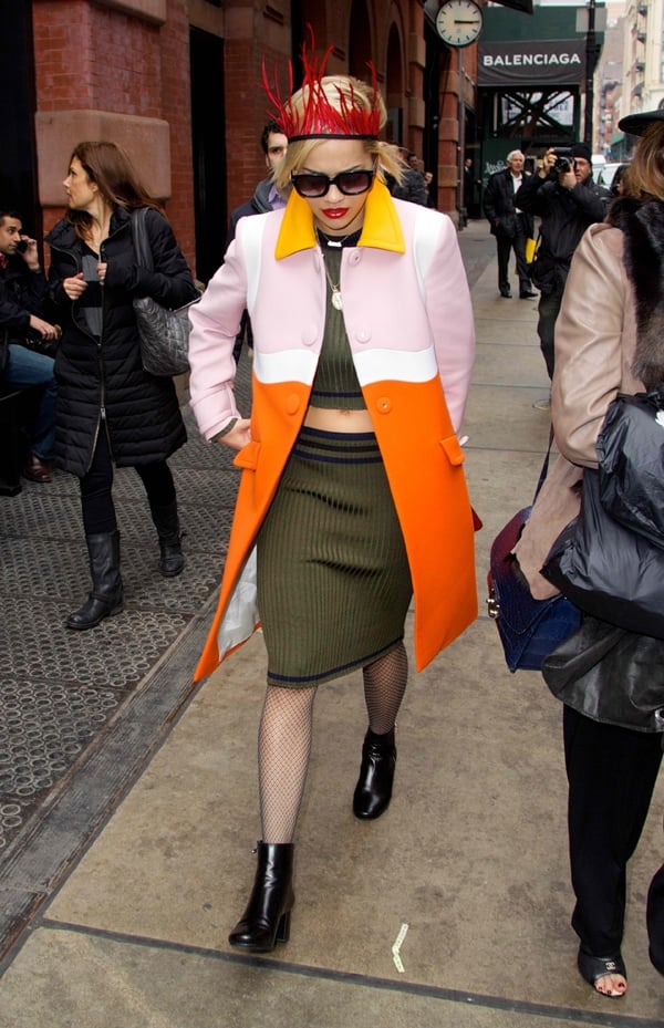 Rita Ora pairs a military green top and matching pencil skirt with a striking red feather headband and a vibrant colorblock coat