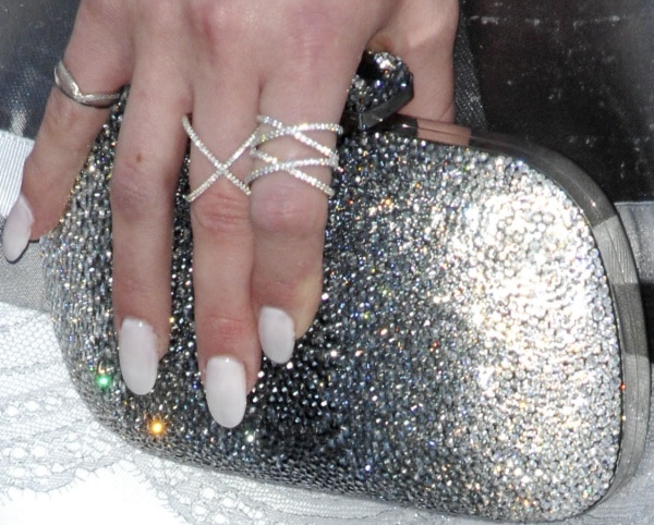 Claudia Lee sports white nails and shows off a selection of sparkling rings while holding a silver embellished clutch