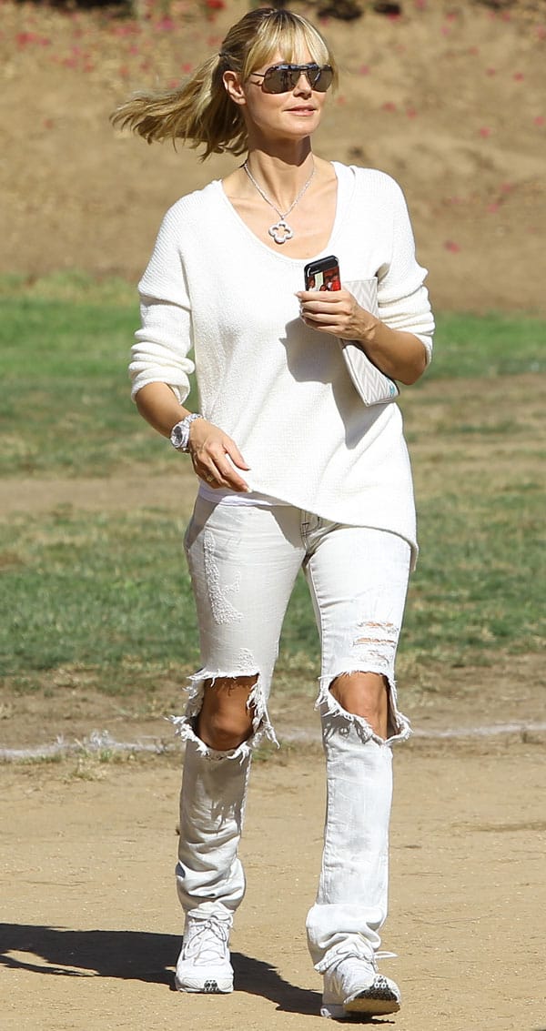 Heidi Klum exudes casual sophistication in ripped white jeans and an asymmetrical hem sweater, accessorized with simple silver jewelry and a clutch for a chic, clean appearance