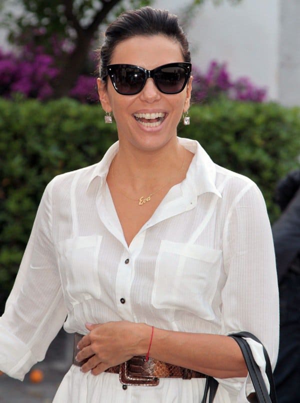 Eva Longoria radiates happiness in a white shirtdress, complemented by a python leather belt, chic sunglasses, and a personalized "Eva" necklace