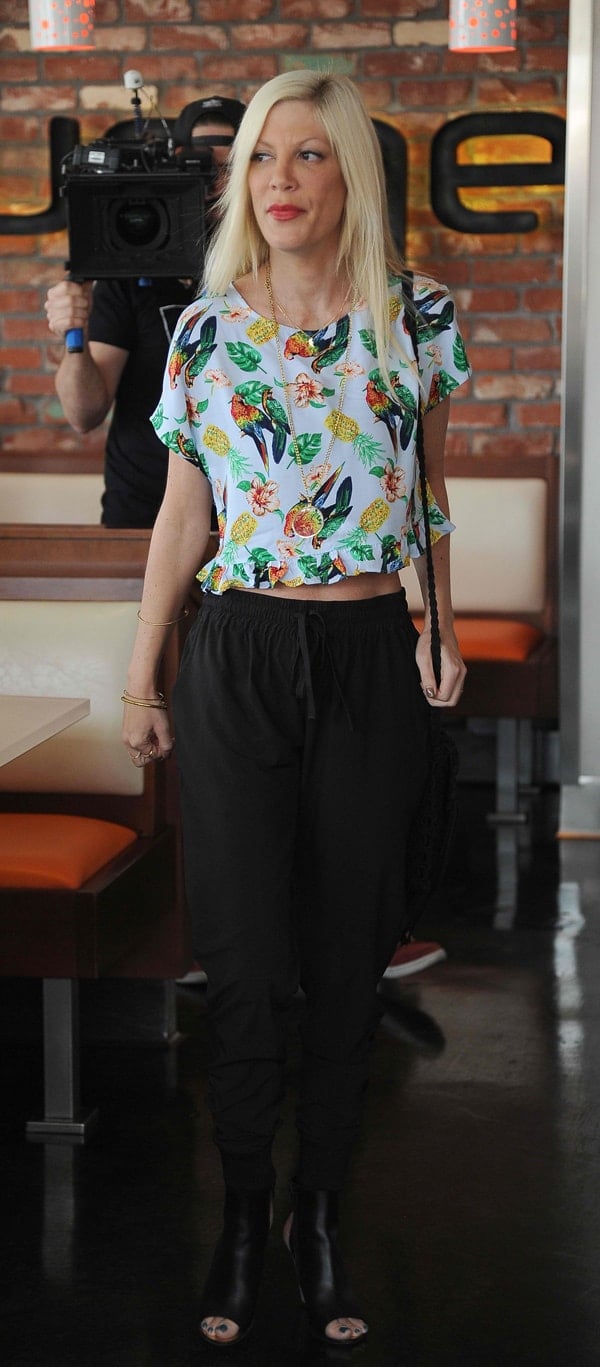 Tori Spelling embraces a playful vibe with a tropical print cropped shirt and black drawstring pants, completed with chic peep-toe booties