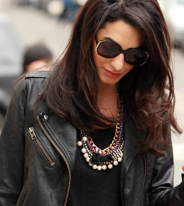 Amal Alamuddin exudes edgy elegance with her stylish ensemble, featuring a sleek black leather biker jacket, oversized dark sunglasses, and a striking statement necklace adorned with colorful stones and pearls