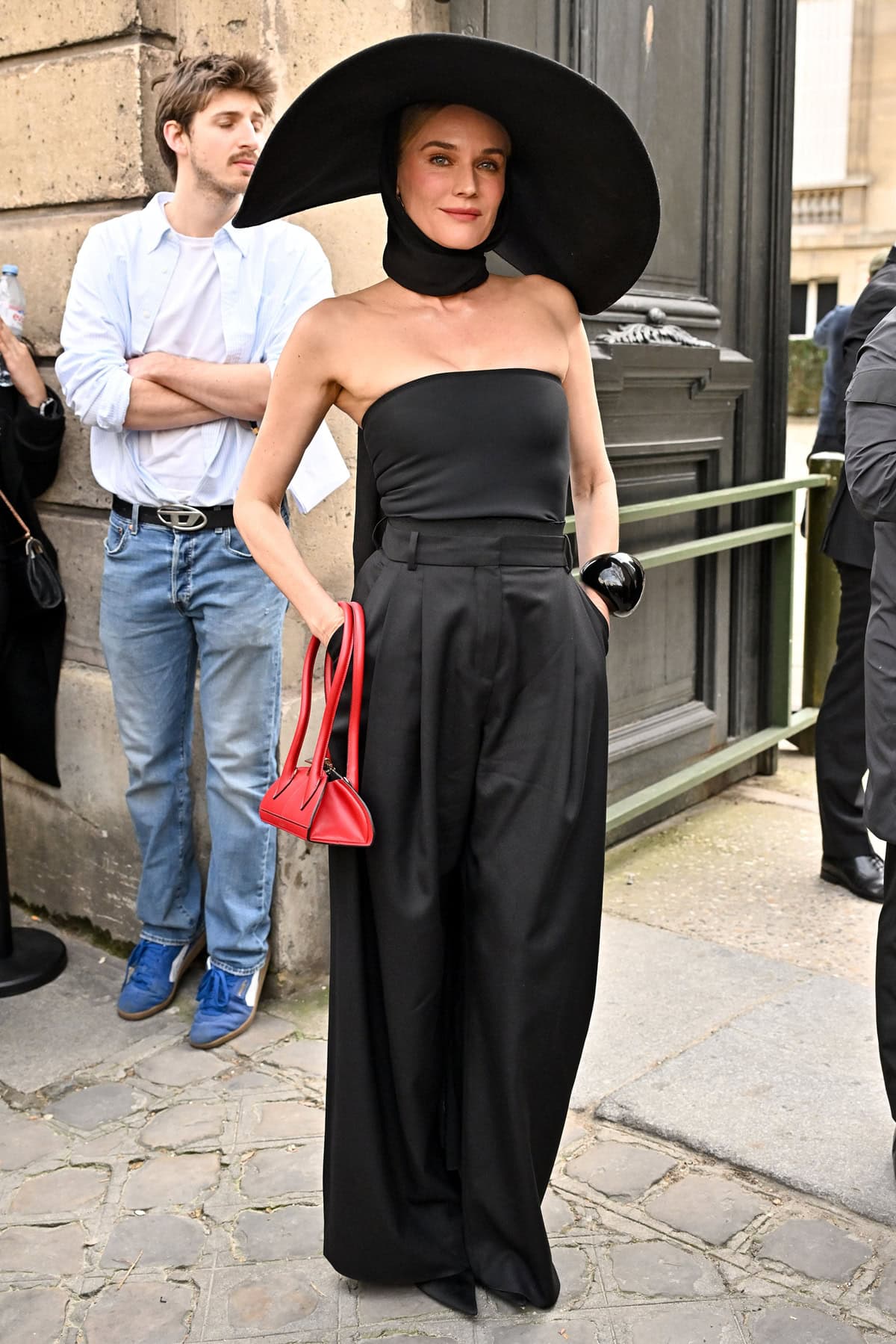 Diane Kruger makes a bold entrance at the Nina Ricci Fall/Winter 2025–2026 show during Paris Fashion Week, wearing a strapless black top, voluminous trousers, a dramatic oversized hat, and a pop of red with a sculptural handbag