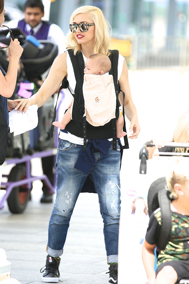 Gwen Stefani effortlessly blends comfort and edge in a white sleeveless top, distressed boyfriend jeans, and Vans Sk8-Hi sneakers, completing her chic mom-on-the-go look with oversized sunglasses and a baby carrier