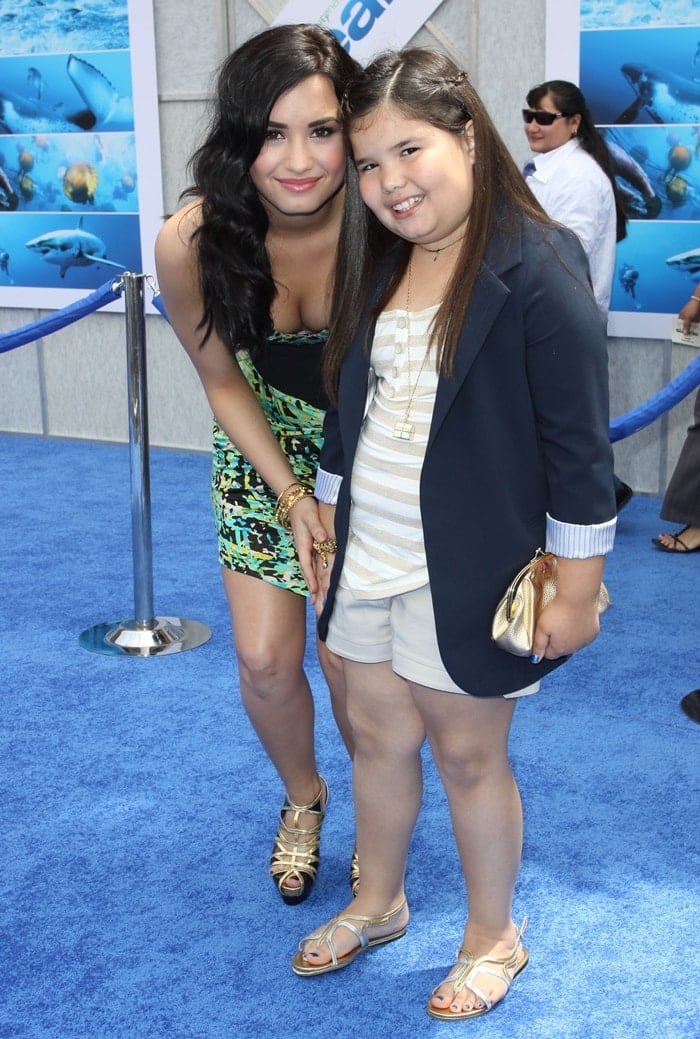 Demi Lovato in strappy gold platform heels and a bold printed mini dress poses with her younger maternal half-sister, actress Madison De La Garza, who wore metallic gold sandals and a navy blazer, at the Oceans Los Angeles Blue Carpet Premiere at the El Capitan Theatre on April 17, 2010, in Hollywood, California