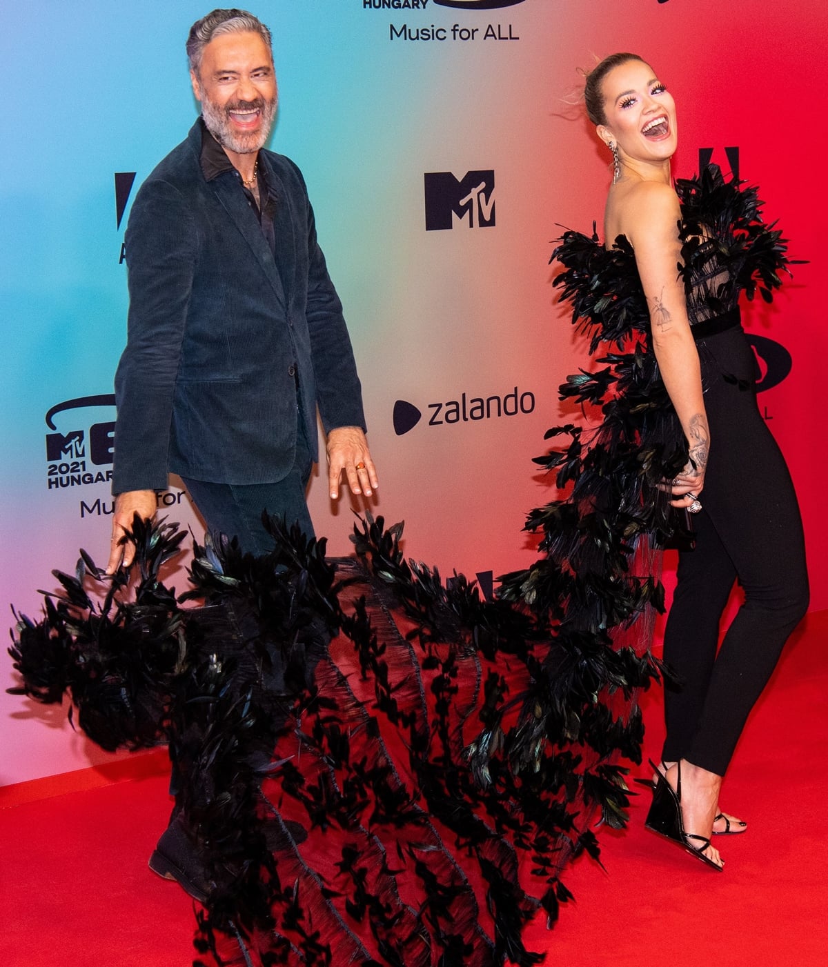 Taika Waititi has fun with Rita Ora's feather train on the red carpet at the Papp Laszlo Budapest Sportarena