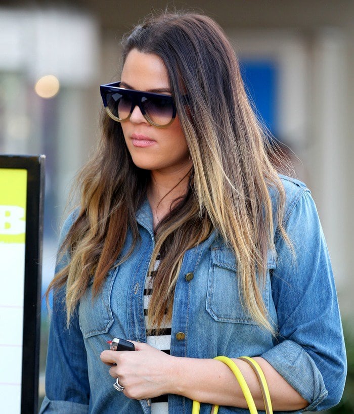 Khloé Kardashian sporting a light-washed denim shirt paired with designer shades, nailing casual glam on a sunny day out